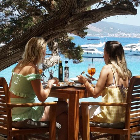 Pair of female charter guests dining at Cala Bassa overlooking anchored motor yacht charters