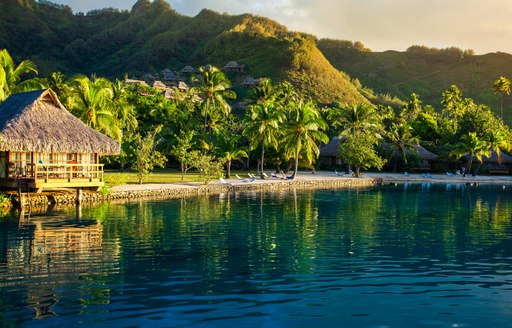 overwater hut faces out onto the water surrounded by palm trees as the sun sets