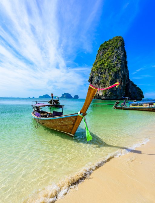 long tailed boats are pulled up onto a golden beach lapped by green waters with a limestone rocky outcrop in the distance